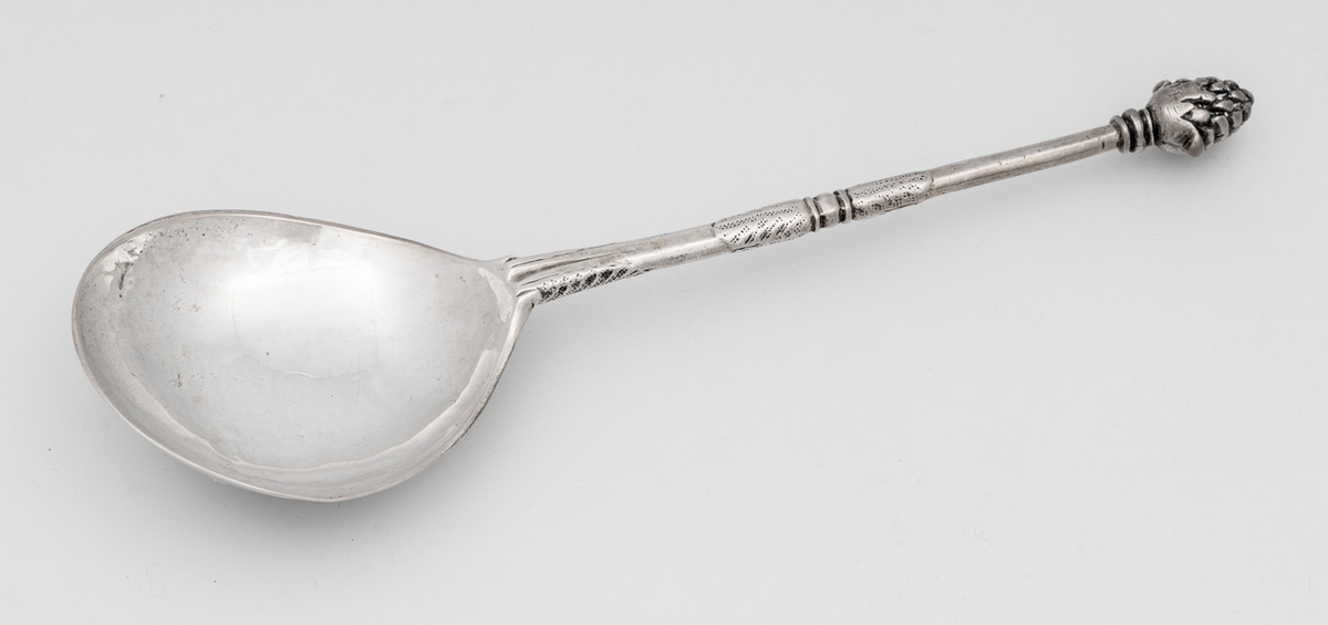 Skje i sølv med dråpeformet blad. Langt, rundt skaft med nedsenket rille nederst med bladornamentikk, et profilert midtparti, samt en drueklase i avslutningen. Baksiden av bladet er med krans og initialer.