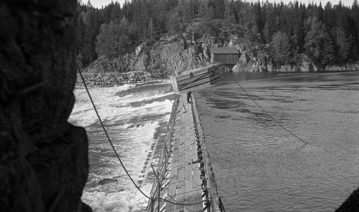 Bygging av såkalt fermettedam ved Mørkfoss i nedre del av Glomma høsten 1914.  Dette er en forholdsvis lett, men robust nåledamtype, der nålene holdes på plass av stålbukker som er hengslet til botnelementer på en måte som gjør det overkommelig å demontere dem om det skulle være påkrevet i reparasjonsøyemed.  Fotografiet er på tvers av vassdraget med damkonstruksjonen sentralt i bildeflata.  Øverst på "baksida" av de nevnte bukkene var det montert gangbruer, som fløterne kunne arbeide fra når dammen skulle settes eller sprettes.  Midt i elveløpet var det en åpning, hvor vannet fosset ut.  Over konstruksjonen ser vi ei taubane med løperkatt, som åpenbart ble brukt til å heise på plass damelementer.  Da dette fotografiet ble tatt var det steinkasser, som skulle plasseres i damløpet for å heve vannstanden i Øyeren, som ved lere anledninger hadde sunket under laveste regulerungsmål.  På motsatt side av elva ser vi et berglendt terreng med noe som kan se ut til å ha vært et redskapshus. Dammen på fotografiet ble sannsynligvis demontert vinteren 1924, for fra påfølgende vår var det den da nye dammen ved Solbergfossen som regulerte Øyeren.  

Den første fermettedammen ble konstruert av den franske ingeniøren Poirée i forbindelse med kanaliseringa av Mainz så tidlig som i 1831.  Dette anlegget ved Mørkfoss kan ha vært det første i sitt slag her til lands.  Denne dammen må ha virket som en terskel i vassdraget, i og med at den hevet vannspeilet oppover mot Øyeren, og dermed gjorde det enklere å få tømmeret fra Øyeren ned til Mørkfoss uten at det strandet på steinskjær og grusører, samtidig som det ble mulig å gå ned i dette området med små slepebåter. 

Fra gammelt av var Mørkfoss en vanskelig passasje for aktørene i tømmerfløtinga.  Vassdragsingeniøren Gunnar Sætren beskrev fossen og den effekten det avbildette damanlegget på stedet fikk slik (1904):

«Umiddelbart nedenfor Øieren er Mørkfossen.  Denne bestod oprindelig af tre fald, og da den var meget smal, havdes særdeles høie flomme i Øieren.  Dette forhold er nu betydelig forbedret, efterat der i aarene 1857 til 69 blev udført betydelige sprængningsarbeider til utvidelse af Mørkfossen.  Samtidig opførtes en reguleringsdam ovr Mørkfossen for at hæve lavvandet i Øieren af Hensyn til dampbaadsfarten og flødningen.  Ved disse arbeider er store flomme sænket betydelig ....»