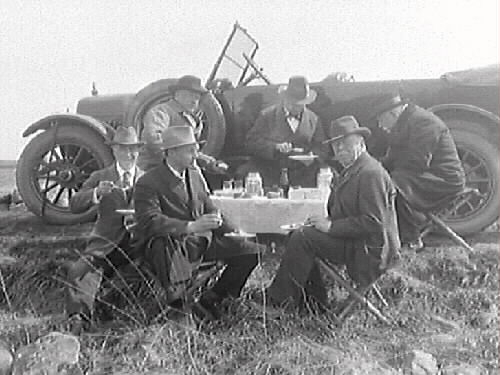 Sex herrar har matpaus vid utgrävningen av Galtabäcksskeppet. En av dem är dr Hallström som inspekterar utgrävningen. Fotofrafen var chef vid Varbergs museum. De sitter i lä bakom en bil.