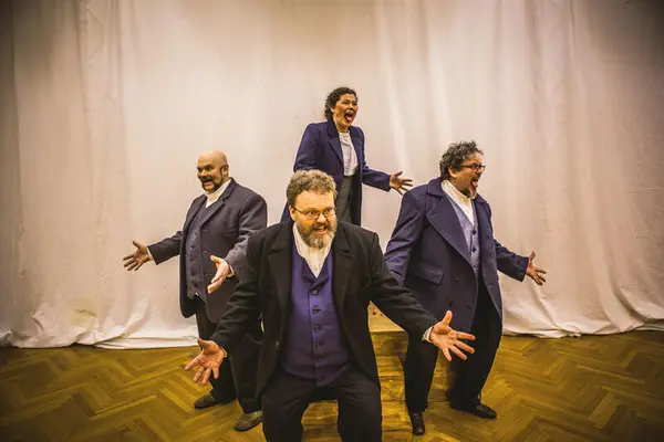 Tre menn og en dame kledd i blå dresser med vester og hvit skjorte. De står i firkantform med spissen ned.