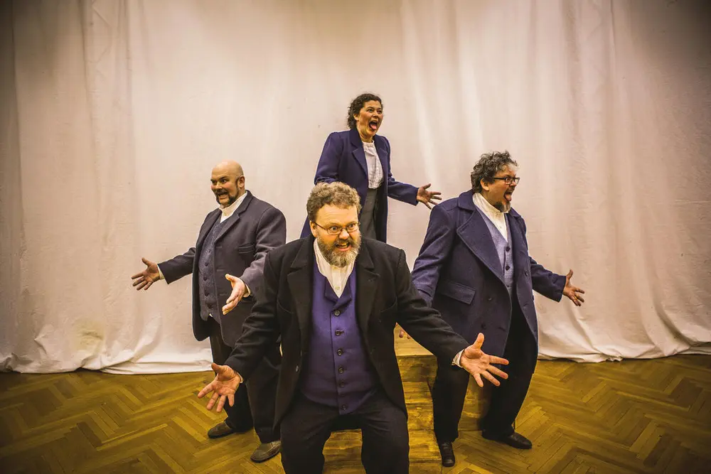 Tre menn og en dame kledd i blå dresser med vester og hvit skjorte. De står i firkantform med spissen ned.