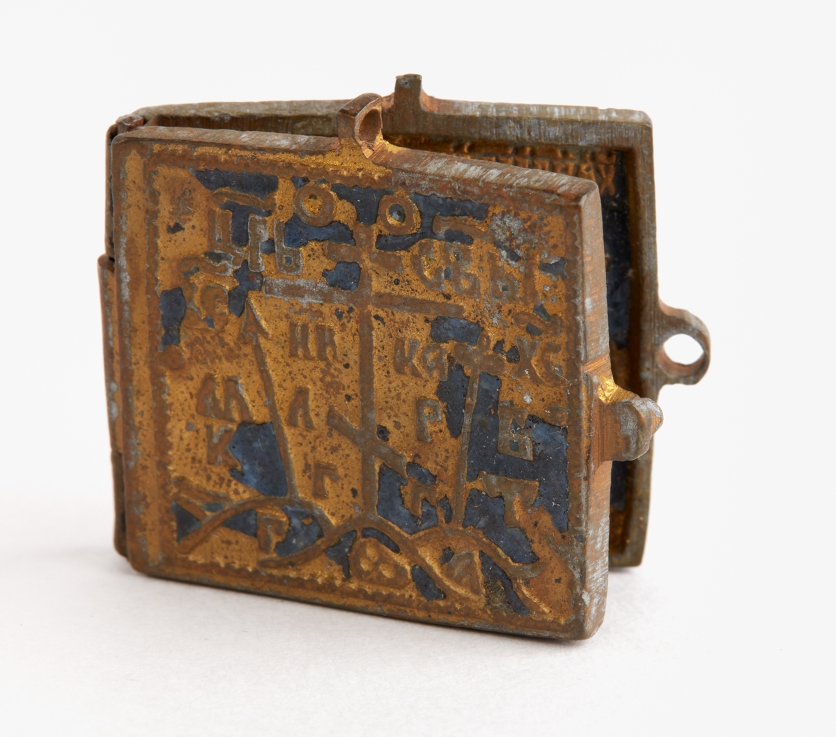 Amulett i förgylld mässing, emalj.

Bredd: 37 mm. 
Höjd: 32 mm. 

Bokform. Yttersidorna med blå emaljbeläggning. Framsidan med grekiska korset samt slavisk inskrift m m: baksidan med ornament. Innersidorna med blå och vit emaljbeläggning; vänster sid; madonnan med barnet, varunder 2 glorieförsedda mansfigurer. Höger sid, i mitten Kristus t v manlig, t h kvinnlig figur i gloria.