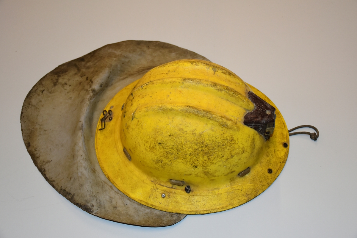 Hjelm i gul plast med fester av metall for lys foran og for ledning til batteri bak . Nakkebeskyttelse av gummiert stoff. Innmaten er laget av skinn, stoffreimer og tau. Hakefeste av lær. Med hakestropp og innmat av plast.