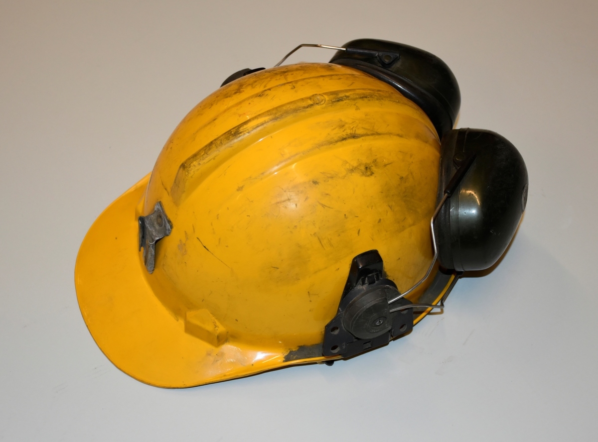 Hjelm i gul plast med fester i metall for lys foran og ledning til batteri bak. Påmontert hørselvern. Innmaten er laget av plast og nylonreimer. Hakerem.