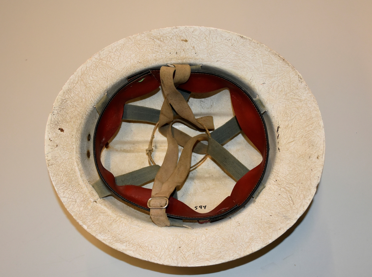 Hjelm i hvit plast med feste av metall for lys foran og feste av lær til ledning bak som gikk til batteri. Innmaten er laget av skinn, stoffreimer og tau. Hakefeste av lær.
