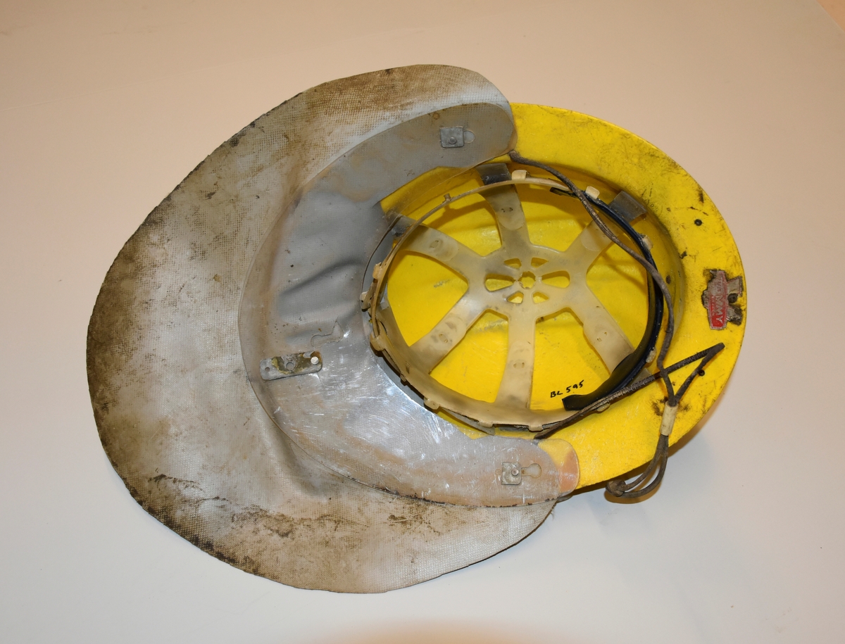 Hjelm i gul plast med fester av metall for lys foran og for ledning til batteri bak . Nakkebeskyttelse av gummiert stoff. Innmaten er laget av skinn, stoffreimer og tau. Hakefeste av lær. Med hakestropp og innmat av plast.