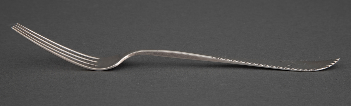Gaffel i sølv. Smalt, langt gaffelblad med fire tinner og skaft som tiltar i bredde mot skaftenden som har spiss avlsutning. Borddekor og gravert navn med bladranke under.