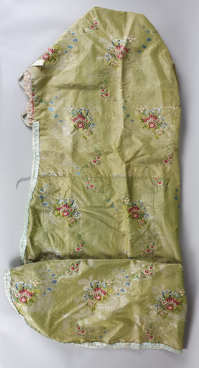 Dåpssveip av brokade, med blomstermønster i rødt, rosa og blått på grønn bunn. Fôret med ubleket lin og med rød silke i hette og nederst. Lyseblå silkekant langs kantene og blonder rundt hetten. Sløyfer og knytebånd.