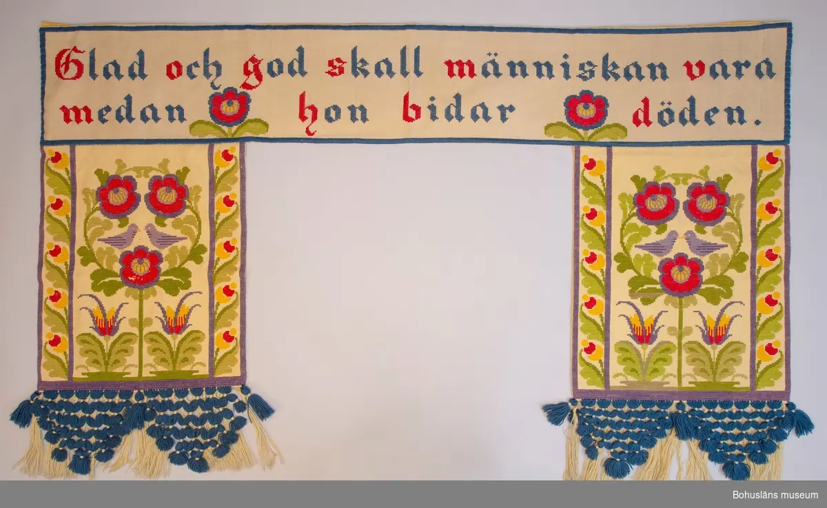 Dörrtextil, rumsavdelare eller draperi i ett stycke. Gul boj broderad i twistsöm i klara färger med stiliserat blomstermotiv med fåglar på sandfärgad botten i jugendstil inspirerad av kurbitsmåleri. 
Överst text i frakturstil med blå bokstäver, inledande bokstaven i rött: 
"God och glad skall människan vara 
medan hon bidar döden".  
150 mm bred knuten yllefrans arrangerad i tofsar i trekantiga knutna former i nederkanten. 
Fodrad med gul bomullssatin. På fodersidan sydd kanal för kraftig gardinstång. 

Gott skick.