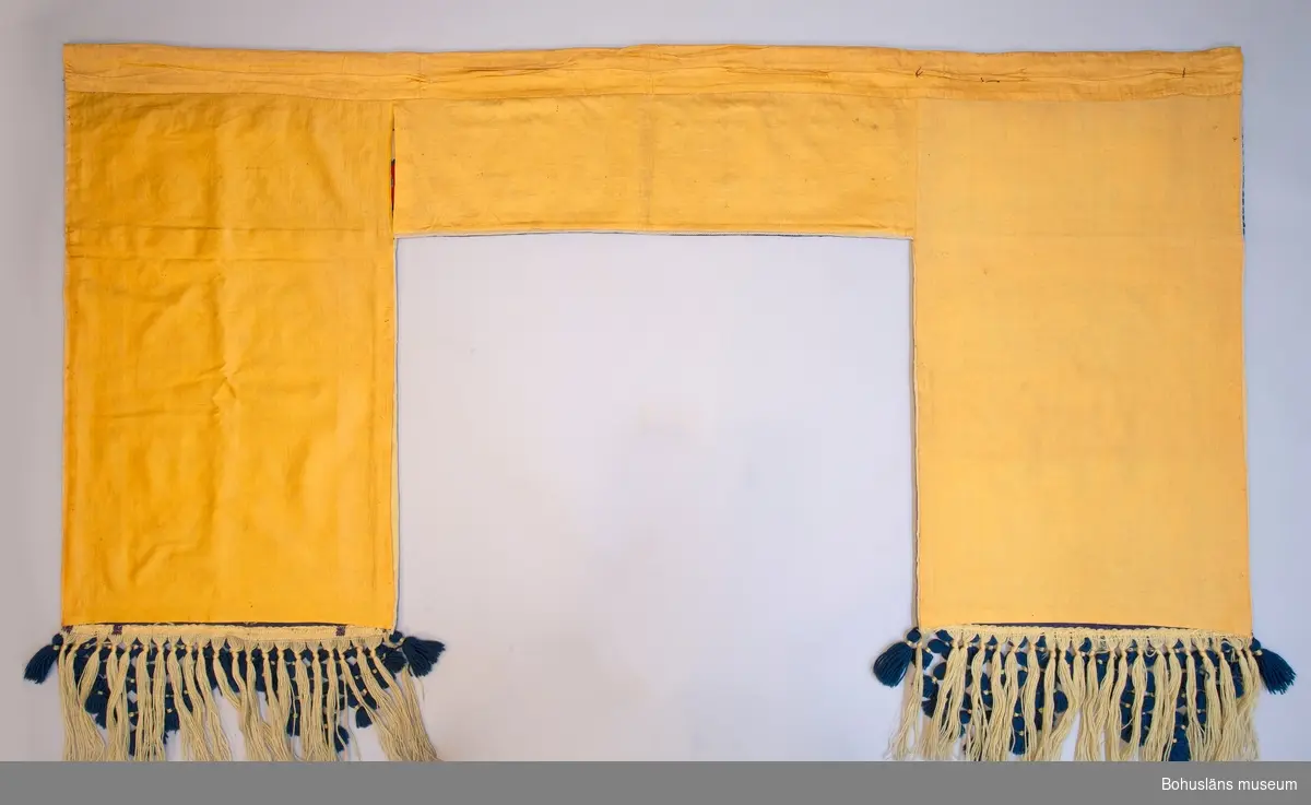 Dörrtextil, rumsavdelare eller draperi i ett stycke. Gul boj broderad i twistsöm i klara färger med stiliserat blomstermotiv med fåglar på sandfärgad botten i jugendstil inspirerad av kurbitsmåleri. 
Överst text i frakturstil med blå bokstäver, inledande bokstaven i rött: 
"God och glad skall människan vara 
medan hon bidar döden".  
150 mm bred knuten yllefrans arrangerad i tofsar i trekantiga knutna former i nederkanten. 
Fodrad med gul bomullssatin. På fodersidan sydd kanal för kraftig gardinstång. 

Gott skick.