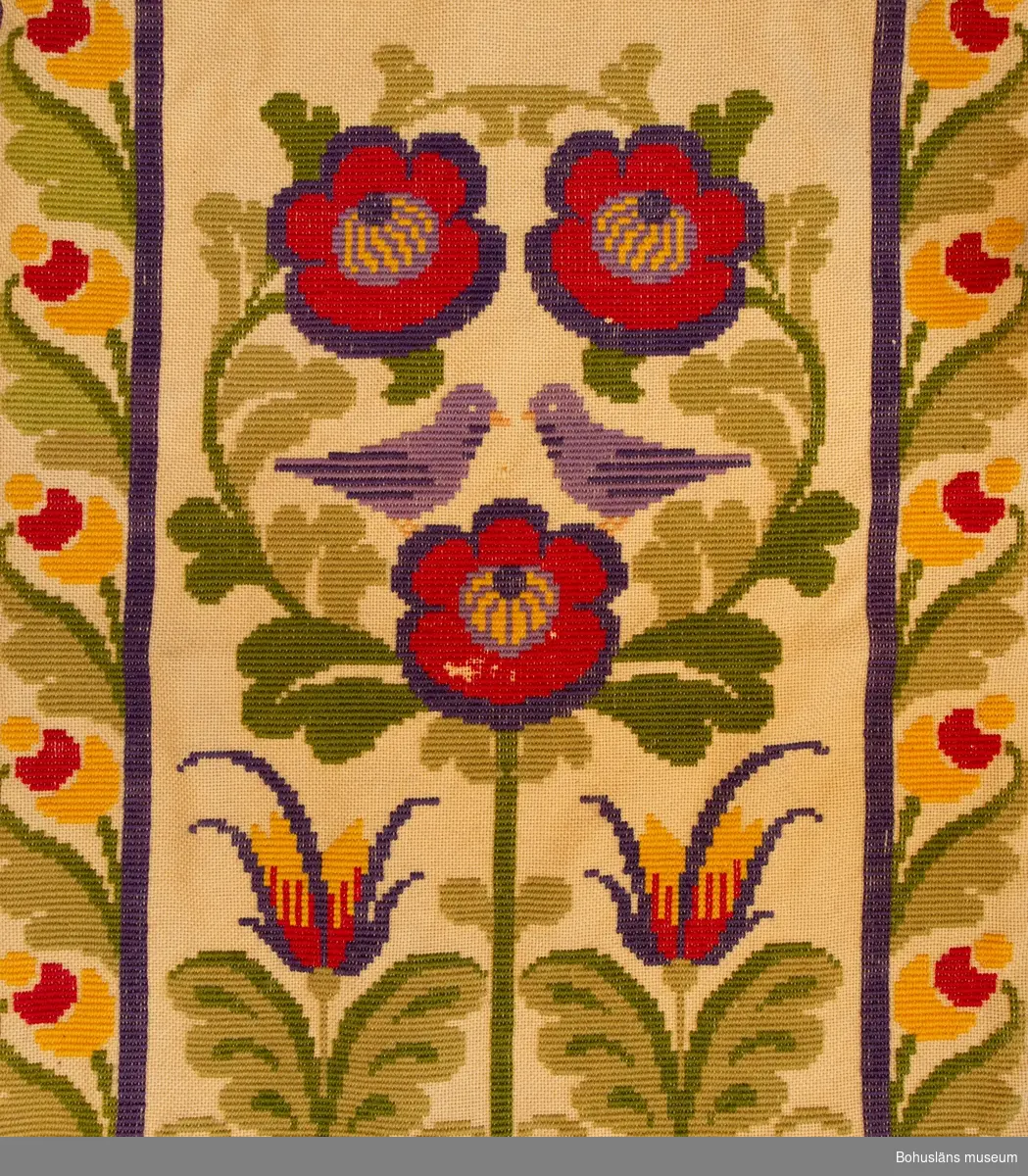 Dörrtextil, rumsavdelare eller draperi i ett stycke. Gul boj broderad i twistsöm i klara färger med stiliserat blomstermotiv med fåglar på sandfärgad botten i jugendstil inspirerad av kurbitsmåleri. 
Överst text i frakturstil med blå bokstäver, inledande bokstaven i rött: 
"God och glad skall människan vara 
medan hon bidar döden".  
150 mm bred knuten yllefrans arrangerad i tofsar i trekantiga knutna former i nederkanten. 
Fodrad med gul bomullssatin. På fodersidan sydd kanal för kraftig gardinstång. 

Gott skick.