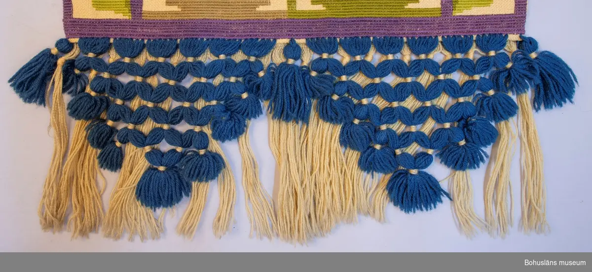 Dörrtextil, rumsavdelare eller draperi i ett stycke. Gul boj broderad i twistsöm i klara färger med stiliserat blomstermotiv med fåglar på sandfärgad botten i jugendstil inspirerad av kurbitsmåleri. 
Överst text i frakturstil med blå bokstäver, inledande bokstaven i rött: 
"God och glad skall människan vara 
medan hon bidar döden".  
150 mm bred knuten yllefrans arrangerad i tofsar i trekantiga knutna former i nederkanten. 
Fodrad med gul bomullssatin. På fodersidan sydd kanal för kraftig gardinstång. 

Gott skick.