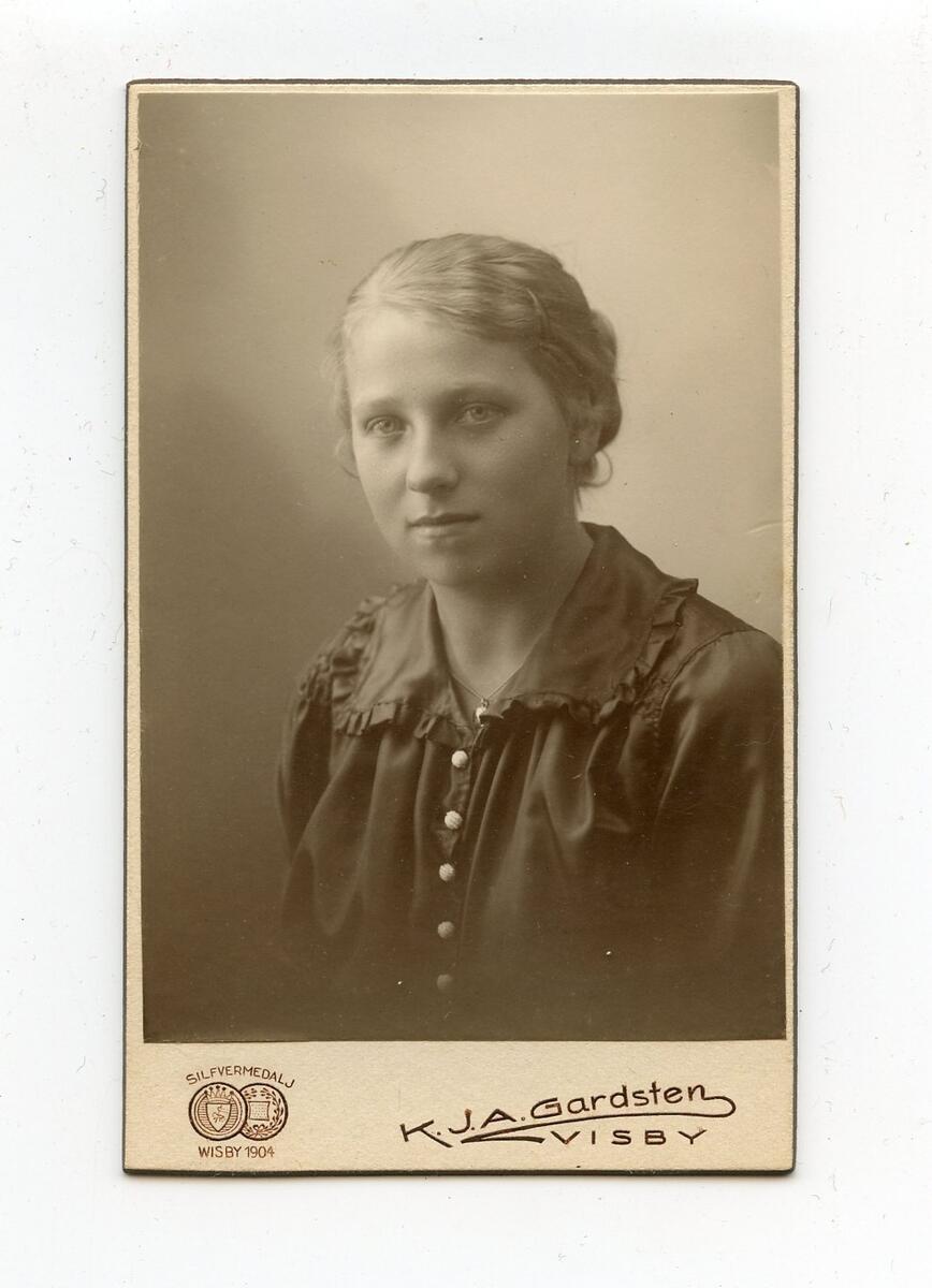 Porträttfotografi i visitkortsformat, tidigt 1900-tal. Bilden föreställer en oidentifierad ung kvinna med ljust hår. Hon är klädd i en mörk blus med ljusa knappar och krage med volang. Fotograf: K.J.A. Gardsten i Visby. Del av en samling porträttfotografier från okänd givare.