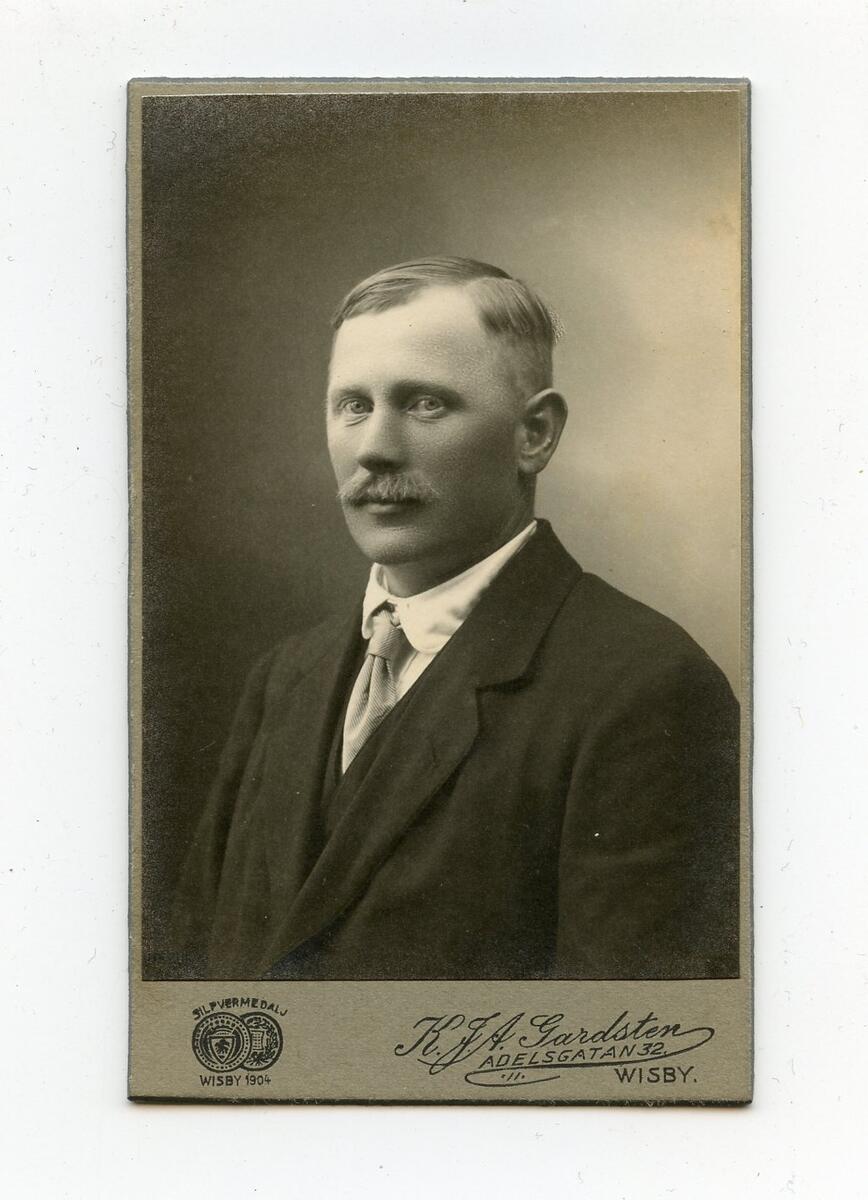 Porträttfotografi i visitkortsformat, tidigt 1900-tal. Bilden föreställer en oidentifierad man med ljust hår och mustasch. Han är klädd i mörk kostym, vit skjorta och ljus slips. Fotograf: K.J.A. Gardsten i Visby. Del av en samling porträttfotografier från okänd givare.