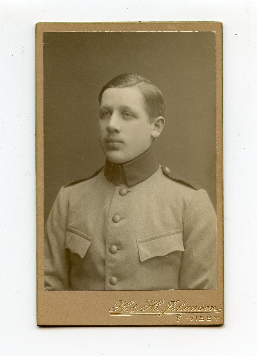 Porträttfotografi i visitkortsformat, tidigt 1900-tal. Bilden föreställer en oidentifierad ung man med ljust hår. Han är klädd i militäruniform med mörk krage. Fotograf: H&H Johanson i Visby. Del av en samling porträttfotografier från okänd givare.