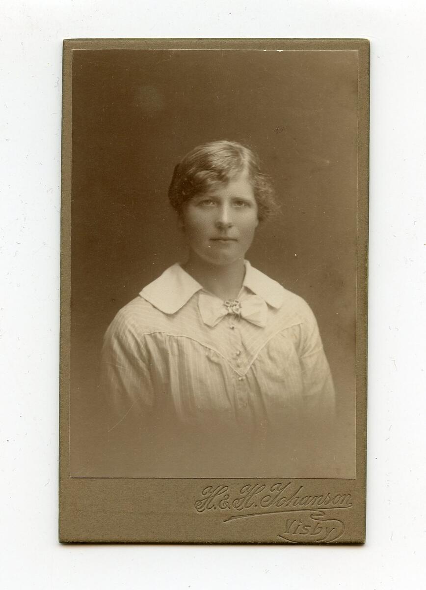 Porträttfotografi i visitkortsformat, tidigt 1900-tal. Bilden föreställer en oidentifierad ung kvinna med ljust hår. Hon är klädd i en ljus, randig blus med krage och rosett. Fotograf: H&H Johanson i Visby. Del av en samling porträttfotografier från okänd givare.