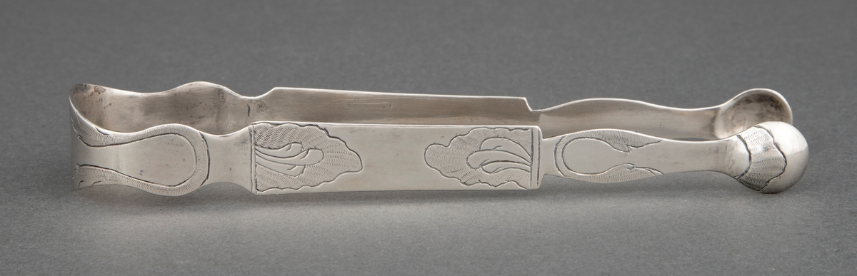 Sukkerklype i sølv. Pinsettform med to små skjeblad ytterst. Hele klypen har profilert randkontur. Enkel, gravert bladdekor over hele klypen.