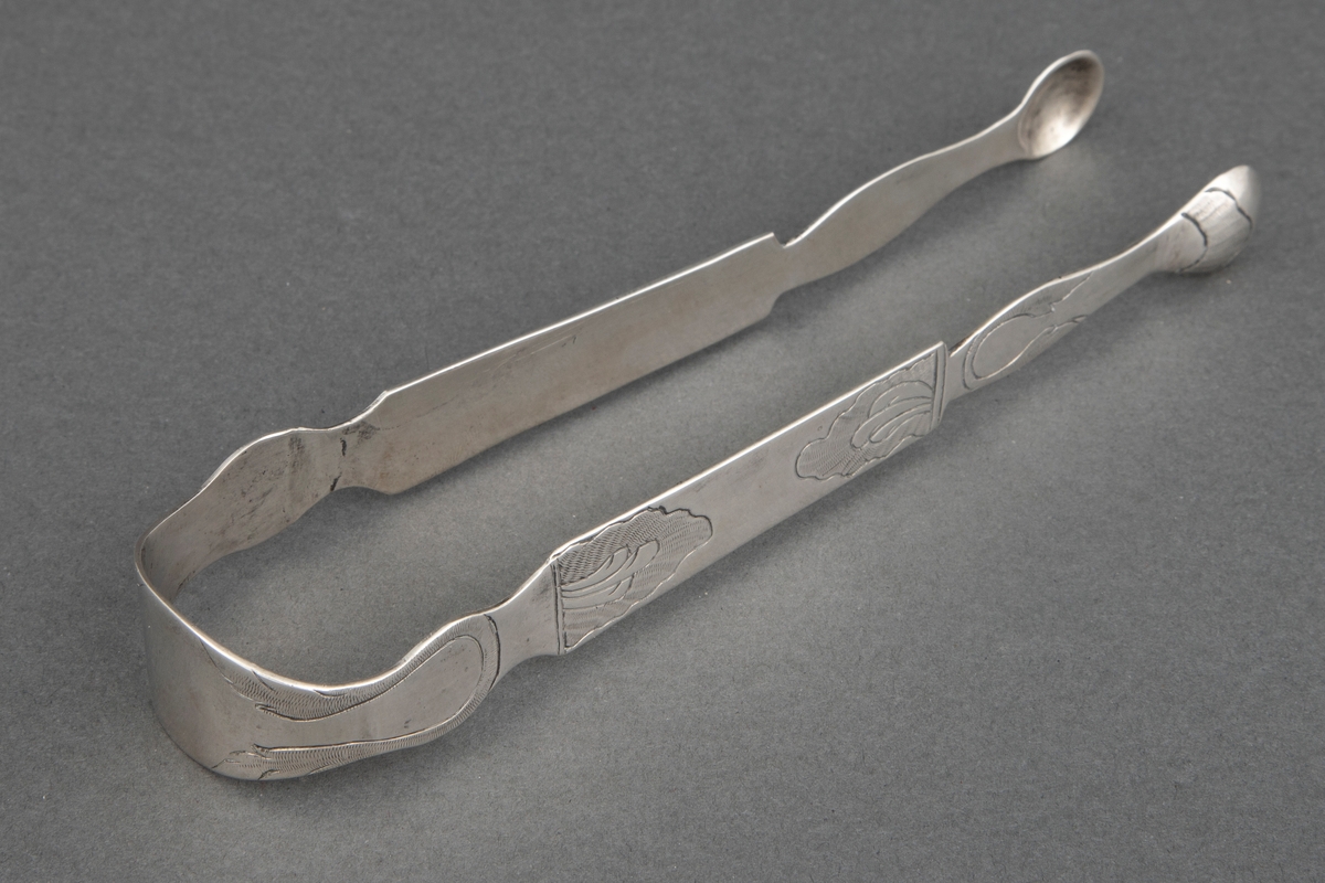 Sukkerklype i sølv. Pinsettform med to små skjeblad ytterst. Hele klypen har profilert randkontur. Enkel, gravert bladdekor over hele klypen.