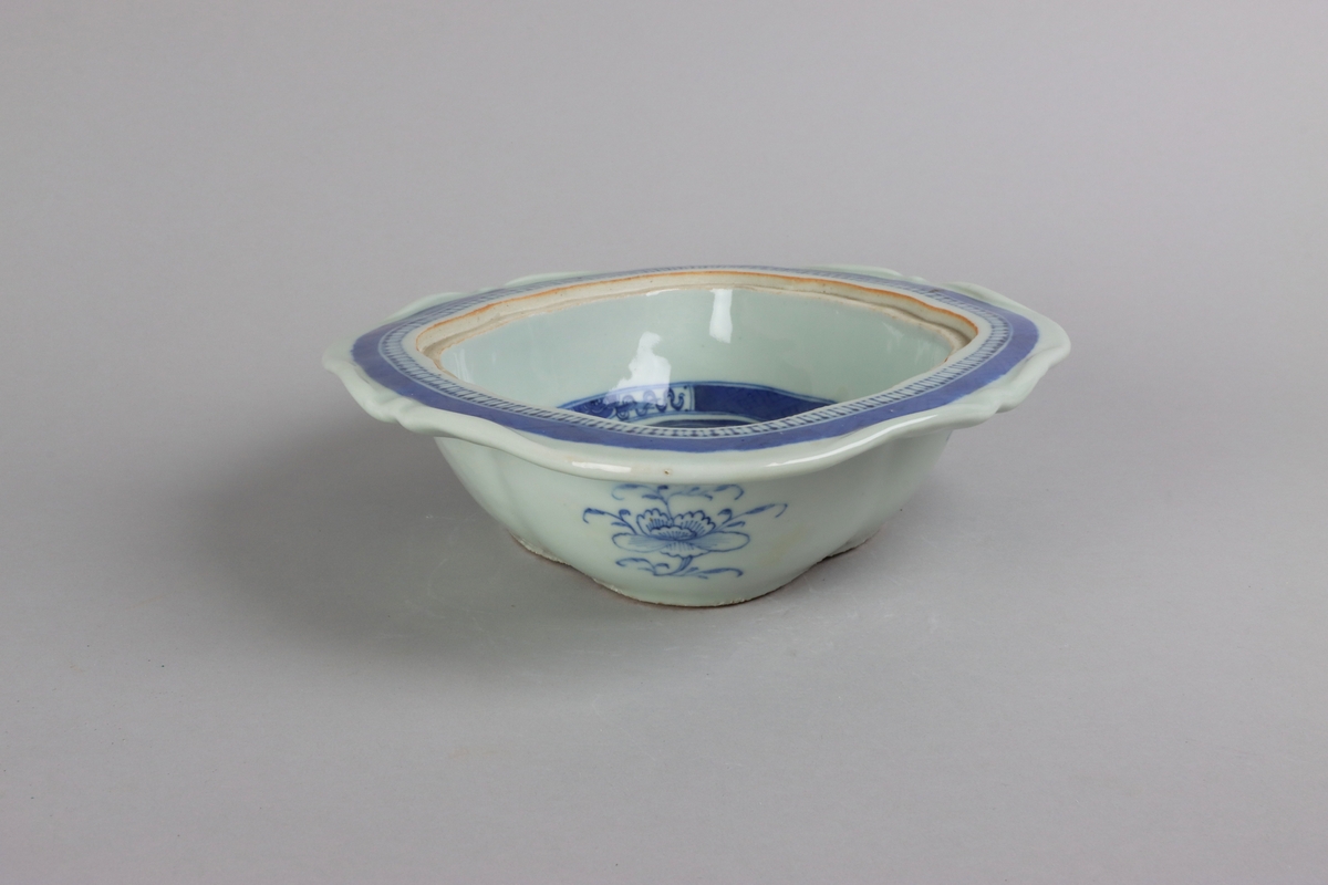Oval broken form with a wide flat rim, decorated with a dark blue border with criss cross pattern. The inside of the dish slightly everted and undecorated. The well surrounded with a border of a criss cross pattern and rectangular reserves filled with symbols of good fortune. Centered in the well landscapes scenes with pagodas, buildings, waters, bridges and gardens. The outside of the dish is decorated with rose branches. All decor in blue underglaze. The base of the dish without glazing.