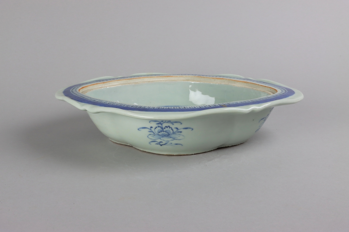 Oval broken form with a wide flat rim, decorated with a dark blue border with criss cross pattern. The inside of the dish slightly everted and undecorated. The well surrounded with a border of a criss cross pattern and rectangular reserves filled with symbols of good fortune. Centered in the well landscapes scenes with pagodas, buildings, waters, bridges and gardens. The outside of the dish is decorated with rose branches. All decor in blue underglaze. The base of the dish without glazing.