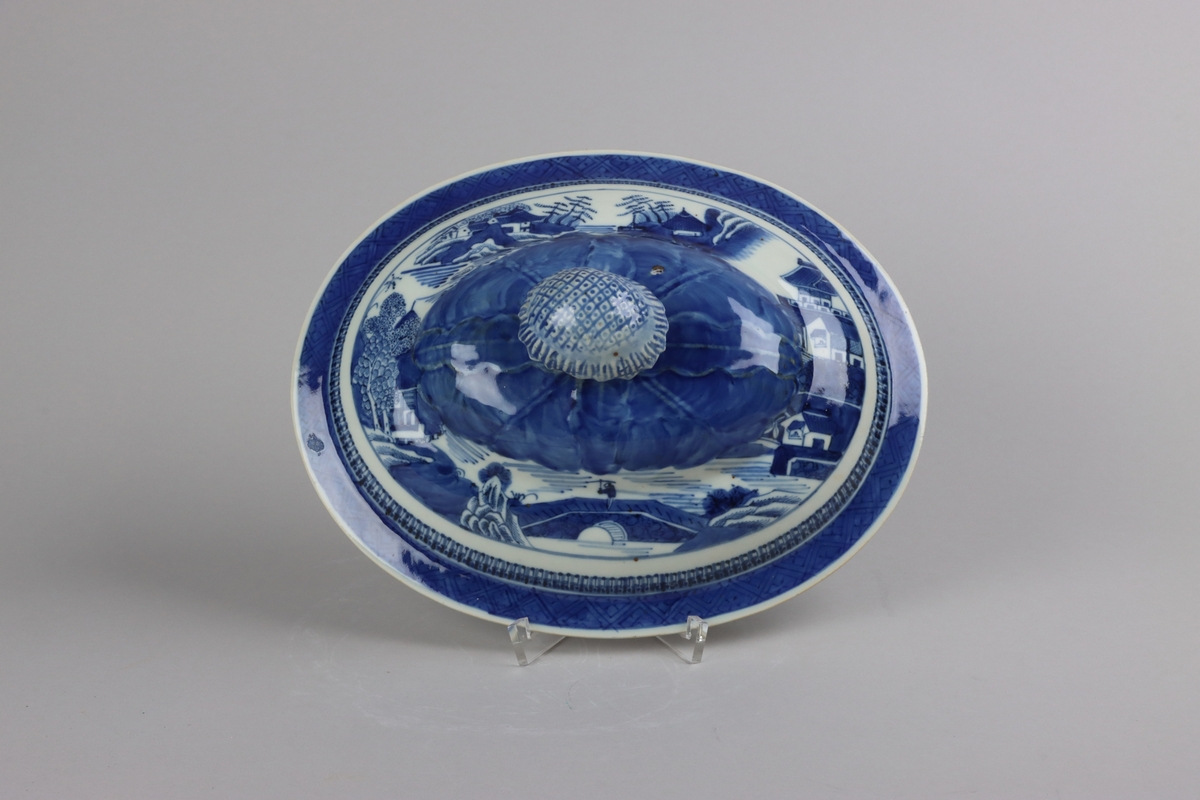 Oval shaped and slightly domed with knob in form of a flower head. On the top of lotus leaves and a list of pagoda lanscapes. The edge of the lid decorated with a dark blue  border in a criss cross pattern. All decor in blue underglaze. The inside of the lid is without decorations.