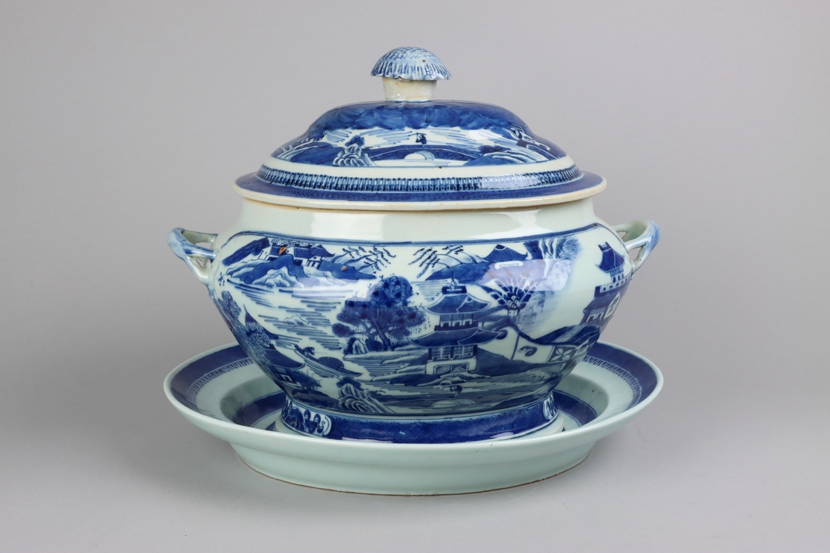 Oval shaped lid with slightly domed form, on top a knob in shape of a flower head. Around the knob lotus leaves, surrounded by a list of pagoda landscapes. The edge of the lid with dark blue border in a criss cross pattern. Oval lobed bowl with angular handles, supported on a central fot. On both sides of the bowl decor of landscape scenes of pagodas, buildings, gardens and waters. Handles in shape of bent tree branches. On the foot a dark blue border with rectangular reserves filled with symbols of good fortune. Oval stand with a lip decorated with dark blue border with criss cross patterns and chrystanthemum flowers. The everted rim is decorated with a dark blue border with a similar pattern as above, but complemented  with oval reserves filled with symbols of fortunes. The well is decorated with landscapes scenes of pagodas, buildings, gardens and bridges. All decor in blue underglaze.