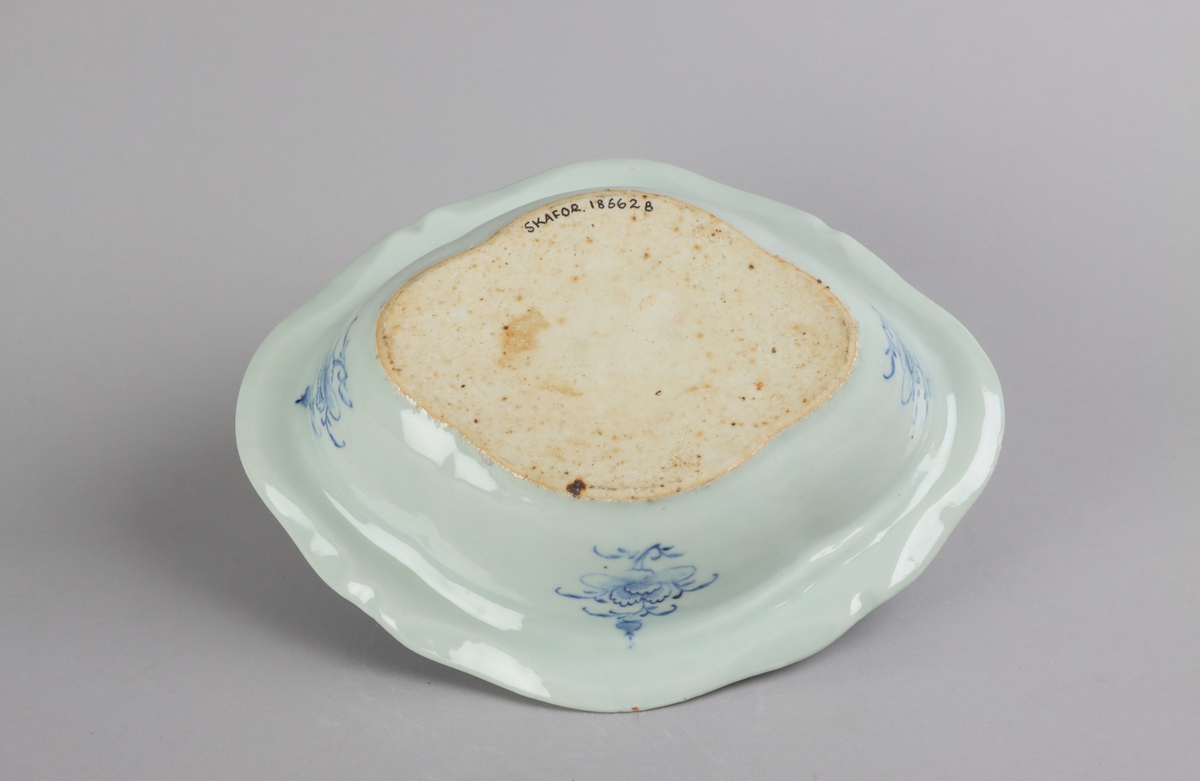 Oval broken form with a wide flat rim, decorated with a dark blue border with criss cross pattern. The inside of the dish slightly everted and undecorated. The well surrounded with a border of a criss cross pattern and rectangular reserves filled with symbols of good fortune. Centered in the well landscapes scenes with pagodas, buildings, waters, bridges and gardens. The outside of the dish is decorated with rose branches. All decor in blue underglaze. The base of the dish without glazing.
