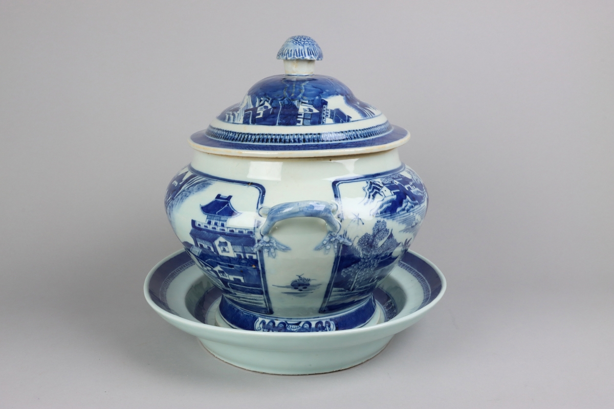 Oval shaped lid with slightly domed form, on top a knob in shape of a flower head. Around the knob lotus leaves, surrounded by a list of pagoda landscapes. The edge of the lid with dark blue border in a criss cross pattern. Oval lobed bowl with angular handles, supported on a central fot. On both sides of the bowl decor of landscape scenes of pagodas, buildings, gardens and waters. Handles in shape of bent tree branches. On the foot a dark blue border with rectangular reserves filled with symbols of good fortune. Oval stand with a lip decorated with dark blue border with criss cross patterns and chrystanthemum flowers. The everted rim is decorated with a dark blue border with a similar pattern as above, but complemented  with oval reserves filled with symbols of fortunes. The well is decorated with landscapes scenes of pagodas, buildings, gardens and bridges. All decor in blue underglaze.