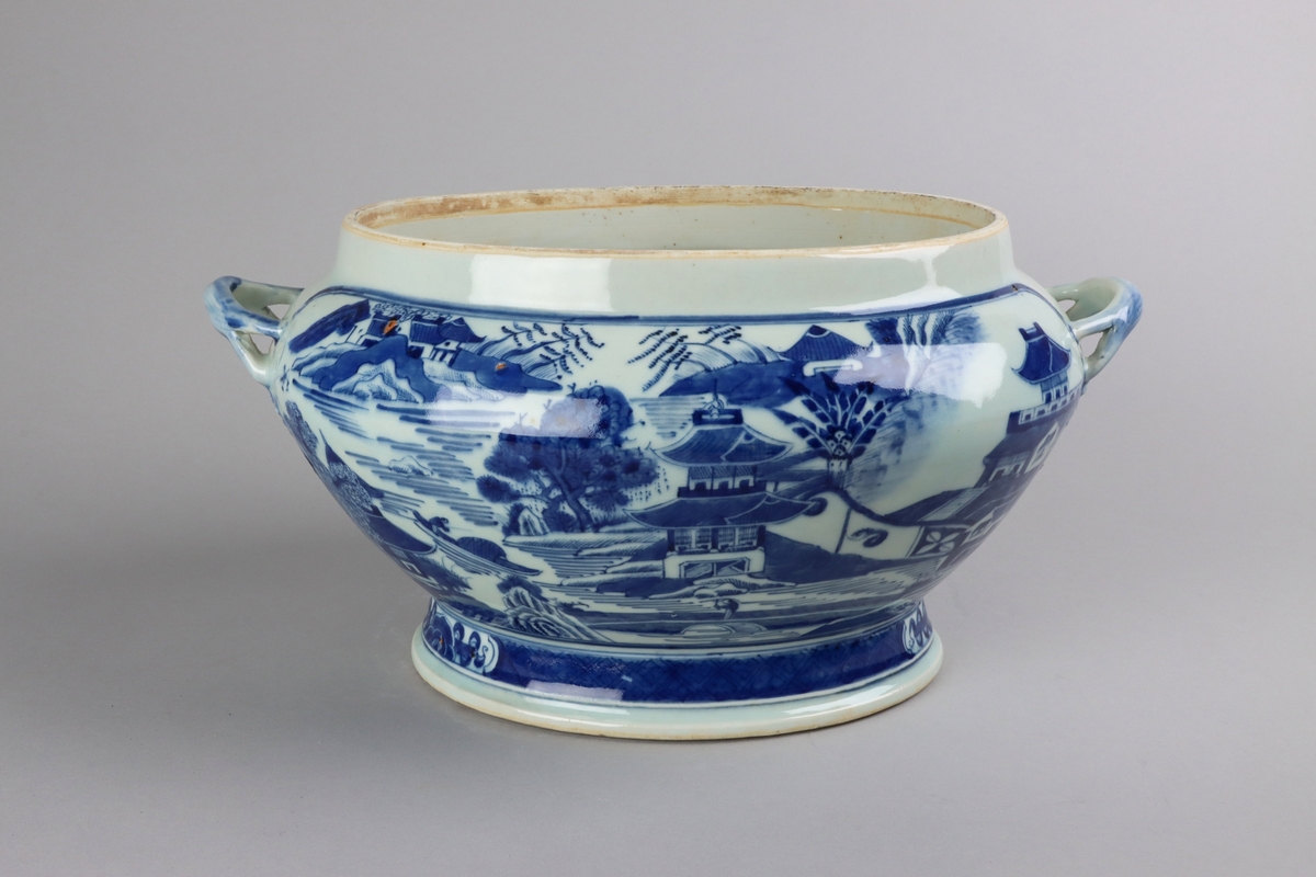 Oval, lobed bowl with angular handles, supported on a central foot. On the both sides decor of landscape scenes of pagodas, buildings, gardens and waters. Handles in shapes of bent tree branches. On the foot a  dark blue border with rectangular reserves filled with symbols of fortunes. All decor in blue underglaze. The base of the bowl  with unglazed ring.