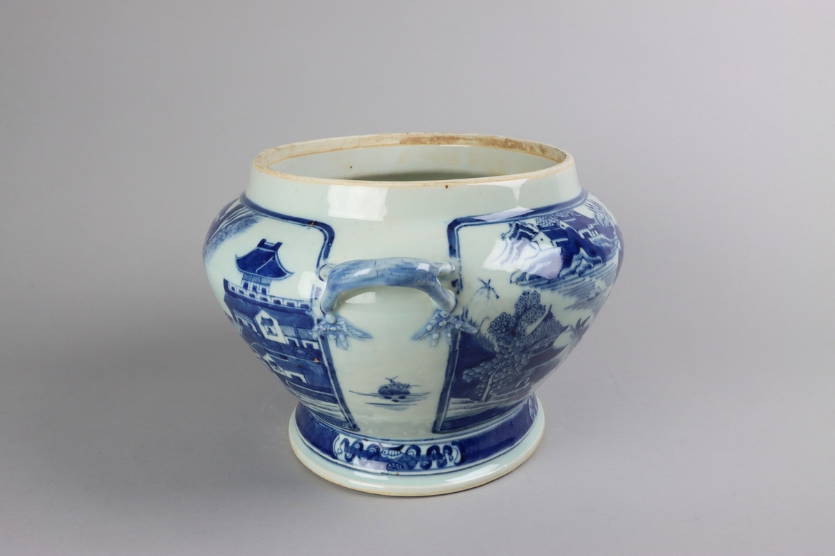 Oval, lobed bowl with angular handles, supported on a central foot. On the both sides decor of landscape scenes of pagodas, buildings, gardens and waters. Handles in shapes of bent tree branches. On the foot a  dark blue border with rectangular reserves filled with symbols of fortunes. All decor in blue underglaze. The base of the bowl  with unglazed ring.
