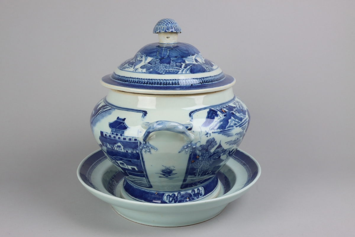 Oval shaped lid with slightly domed form, on top a knob in shape of a flower head. Around the knob lotus leaves, surrounded by a list of pagoda landscapes. The edge of the lid with dark blue border in a criss cross pattern. Oval lobed bowl with angular handles, supported on a central fot. On both sides of the bowl decor of landscape scenes of pagodas, buildings, gardens and waters. Handles in shape of bent tree branches. On the foot a dark blue border with rectangular reserves filled with symbols of good fortune. Oval stand with a lip decorated with dark blue border with criss cross patterns and chrystanthemum flowers. The everted rim is decorated with a dark blue border with a similar pattern as above, but complemented  with oval reserves filled with symbols of fortunes. The well is decorated with landscapes scenes of pagodas, buildings, gardens and bridges. All decor in blue underglaze.