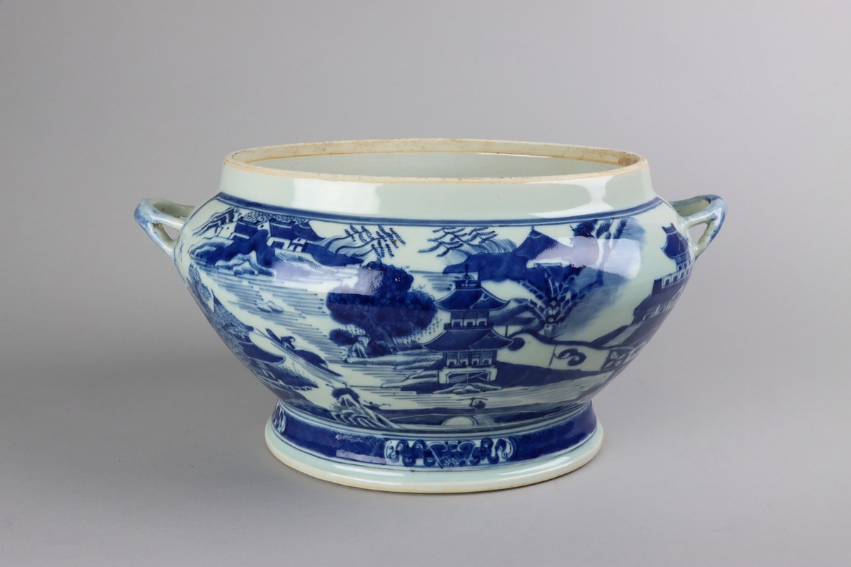 Oval, lobed bowl with angular handles, supported on a central foot. On the both sides decor of landscape scenes of pagodas, buildings, gardens and waters. Handles in shapes of bent tree branches. On the foot a  dark blue border with rectangular reserves filled with symbols of fortunes. All decor in blue underglaze. The base of the bowl  with unglazed ring.