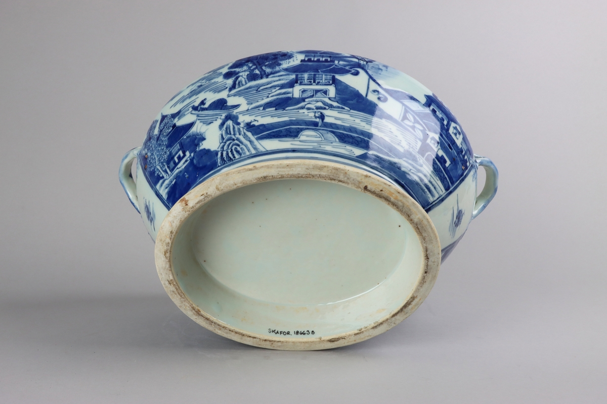Oval, lobed bowl with angular handles, supported on a central foot. On the both sides decor of landscape scenes of pagodas, buildings, gardens and waters. Handles in shapes of bent tree branches. On the foot a  dark blue border with rectangular reserves filled with symbols of fortunes. All decor in blue underglaze. The base of the bowl  with unglazed ring.