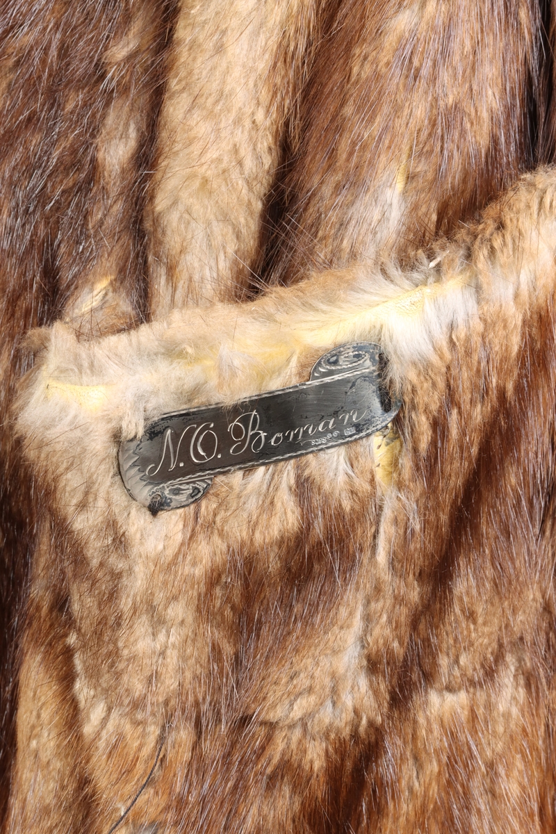 Rock av svart kläde fodrad med brun päls, troligen av bisamråtta. Ärmar fodrade med mörkbrunt fårskinn. Krages slag fodrade med svart persian. Rocken stängs med dubbelknäppning möjlig att knäppa åt båda håll. Sammanlagt sex svarta knappar. Två fickor med lock på framsidan. En innerficka prydd med namnskylt av silver.