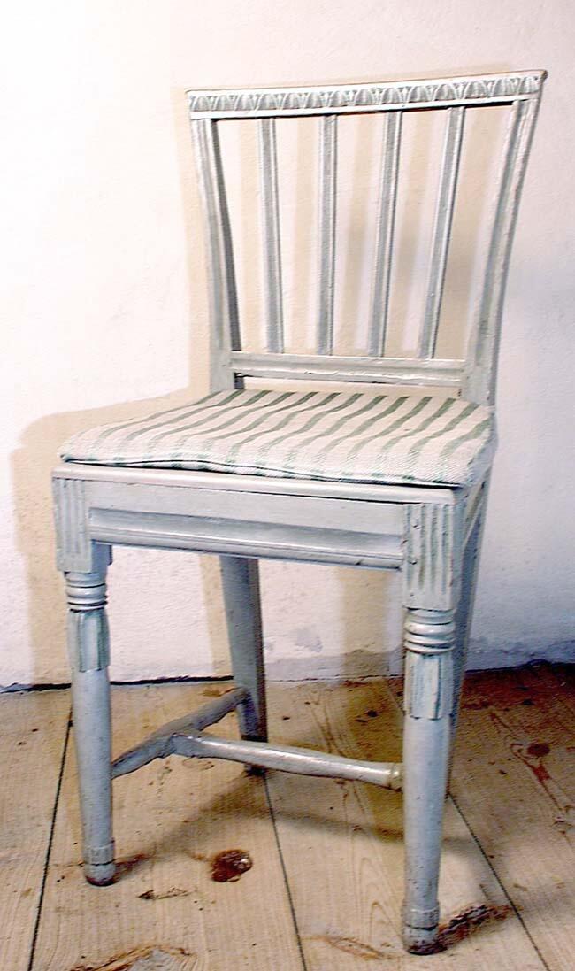 FRÅN GOTLANDS LANDSBYGD. INKÖP; ERIK PERSSON ANTIKHANDEL, VISBY.  H. CA. 89.5 CM, BR. FRAM CA. 42 CM, DJUP 36,5 CM, SITTHÖJD 47 CM.  FURU, BJÖRK, JÄRN, LIN, BOMULL.  2 ST STOLAR ( LIKADANA ).   AV FURU, MED krÖN OCH TVÄRBAND AV BJÖRK. GENOMBRUTEN RYGG, VERTIKALT LÄTT BAKÅTSVÄNGD. SIDOSTOLPAR UTÅTBÖJDA, EN BRED KANNELYR. krÖNET FÖRSETT MED NAGELSNITT. FYRA SPJÄLOR. TVÄRBAND NERTILL MED BRED KANNELYR. FASTS SITS AV FURU, FASTSPIKAD. SARG MED FYRKANTIGA HÖRN. BENENS FÖRLÄNGNING FÖRSEDD MED KANNELYRER. PROFILERAD KANTLIST OCH HÅLKÄL PÅ ALLA FYRA SIDORNA. FRAMBEN RUNDA, MED RINGAR. VID ANSATSEN NEDÅTRIKTAD, STILISERAD BLADHOLK, LÄTT BALUSTERFORMADE, AVSLUTAS MED RÄFFLAD RING OCH KORT BALUSTER.  BAKBENEN FYRKANTIGA, NEDÅT AVSMALNANDE. MÅLADE I PÄRLGRÅTT MED DRAGNING ÅT GRÖNT. LÖS DYNA, TYG, SE PS. 1173. FASTKNUTEN VID RYGGENS SIDOSTOLPAR. NÅGOT MASKÄTNA. ( SITS OCH DYNOR NYPÅSATTA. A. BLOCK ). ( NEDRE KAMMAREN, EN STOL FRAMFÖR PS. 1385, EN FRAMFÖR FÖNSTRET ).