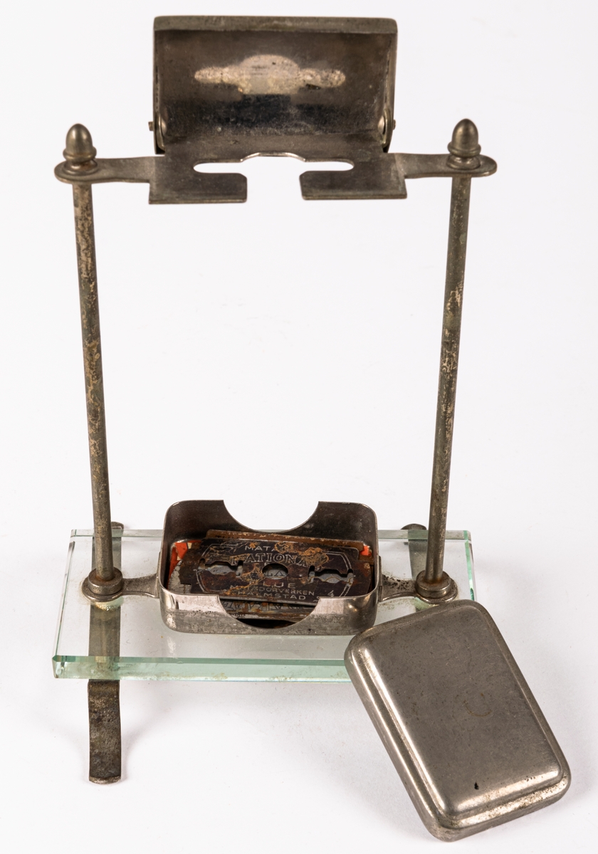 Ställ för rakhyvel. Ställning, skydd för rakhyvel och ask för rakblad i metall. På benen i metall en rektangulär glasskiva på vilken rakbladsasken står. 12 rakblad ingår. Rakbladen märkta SWING, DRABANT, MATADOR 25, GILETTE och MATADOR NATIONAL BLÅ.