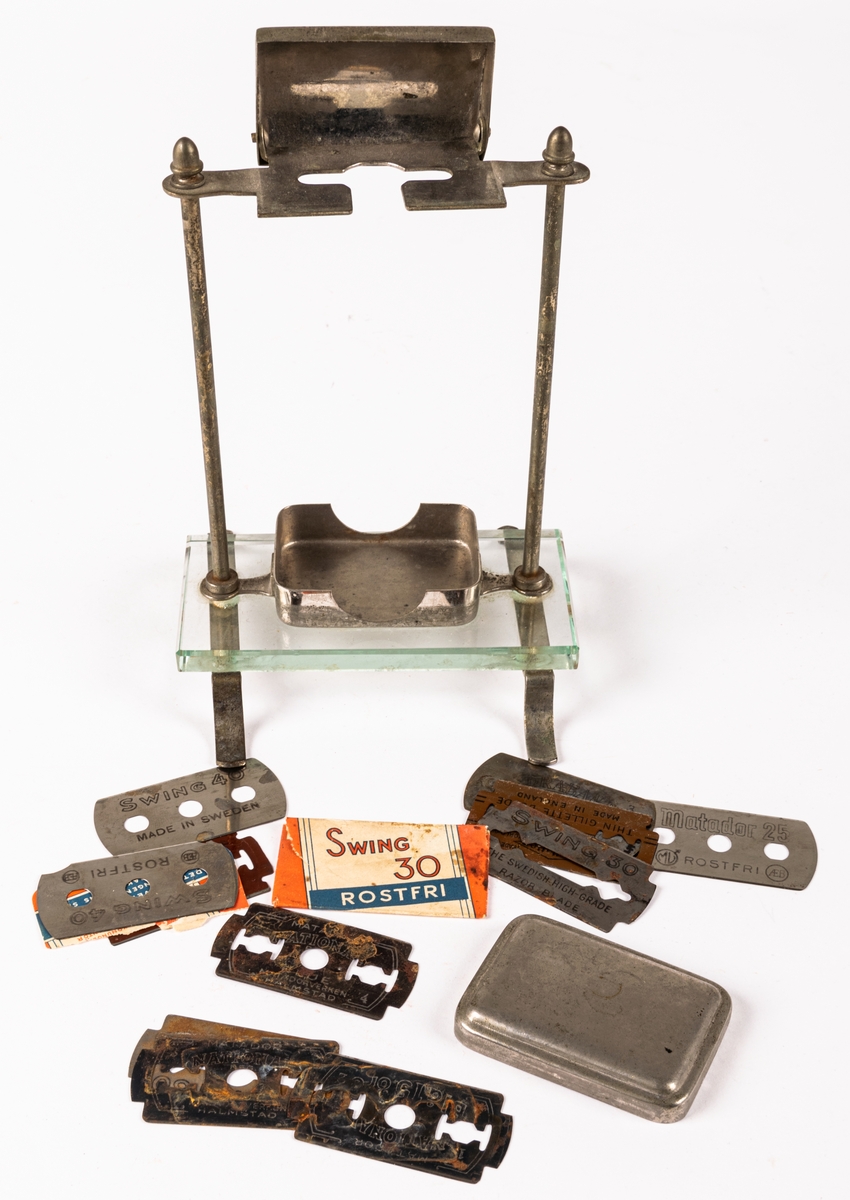 Ställ för rakhyvel. Ställning, skydd för rakhyvel och ask för rakblad i metall. På benen i metall en rektangulär glasskiva på vilken rakbladsasken står. 12 rakblad ingår. Rakbladen märkta SWING, DRABANT, MATADOR 25, GILETTE och MATADOR NATIONAL BLÅ.