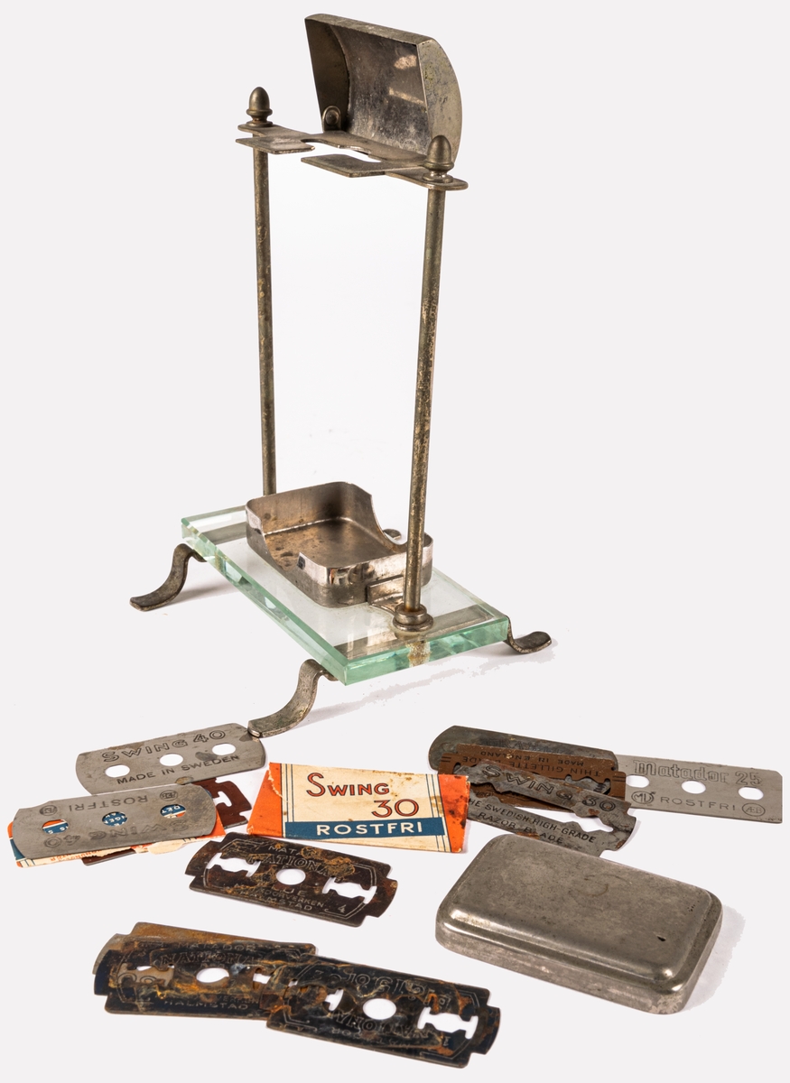 Ställ för rakhyvel. Ställning, skydd för rakhyvel och ask för rakblad i metall. På benen i metall en rektangulär glasskiva på vilken rakbladsasken står. 12 rakblad ingår. Rakbladen märkta SWING, DRABANT, MATADOR 25, GILETTE och MATADOR NATIONAL BLÅ.