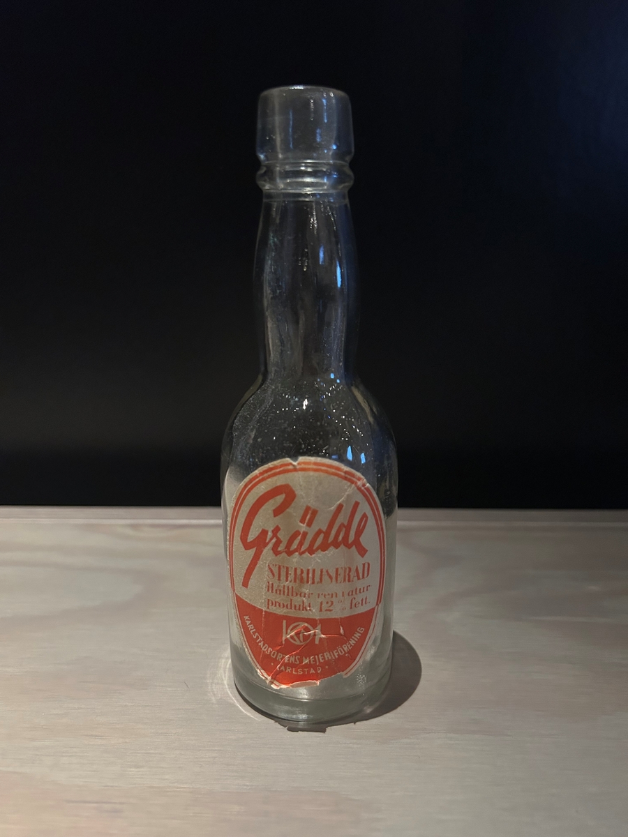 Flaska av ofärgat glas för förvaring av grädde. Hög smal hals. Kapsyl saknas. Oval pappersetikett i gul och orange med text: "Grädde steriliserad Hållbar ren naturprodukt 12 % fett" i det gula fältet, och "Karlstadsortens mejeriförening Karlstad" samt "KM" med en cirkel omkring, i det orangea fältet.