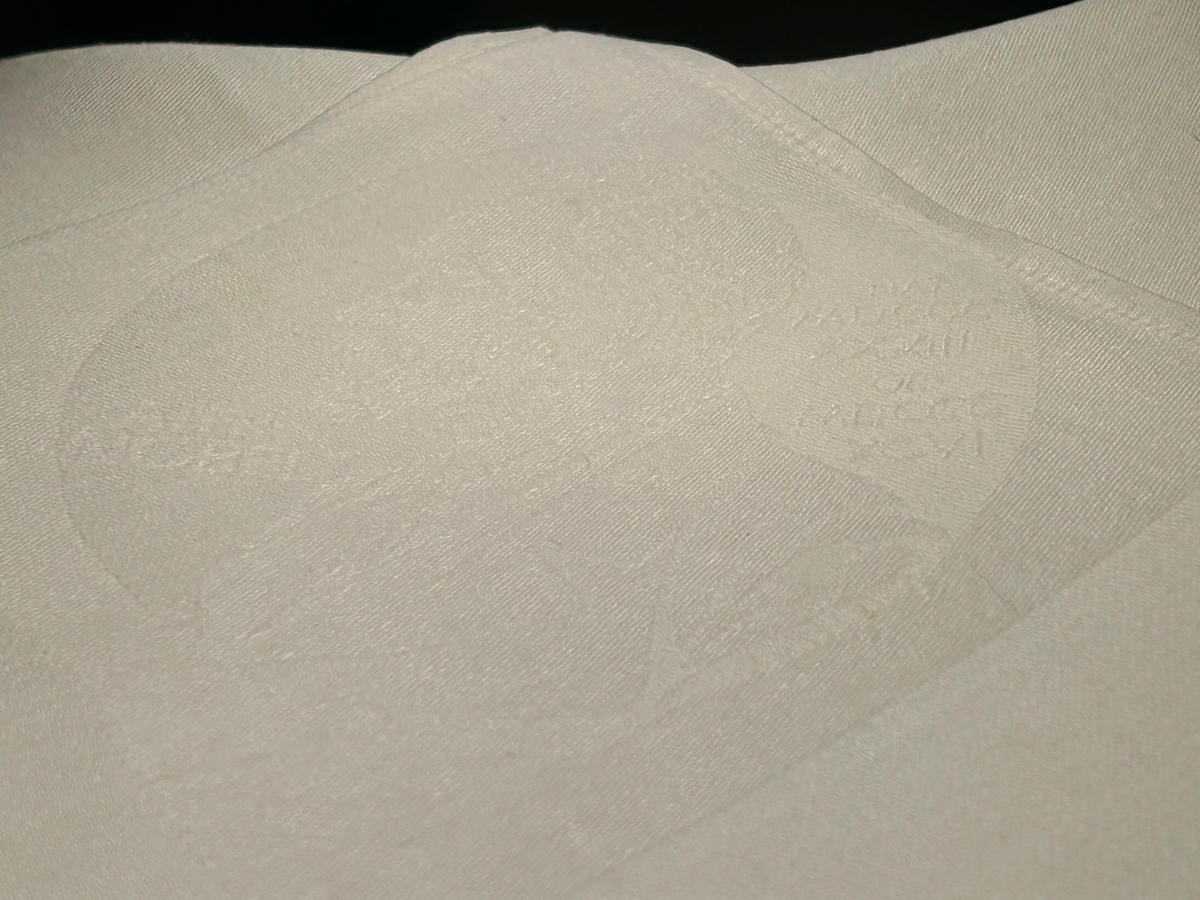 Nobelservetten med emblem i vitt, tunt, lingarn. Det vävda rutmönstret blir synligt genom att ljuset slår olika från varp- och väftytorna. Nobelemblemet är placerat i ett av servettens hörn. En produktetikett i papper är fästad i servetten.