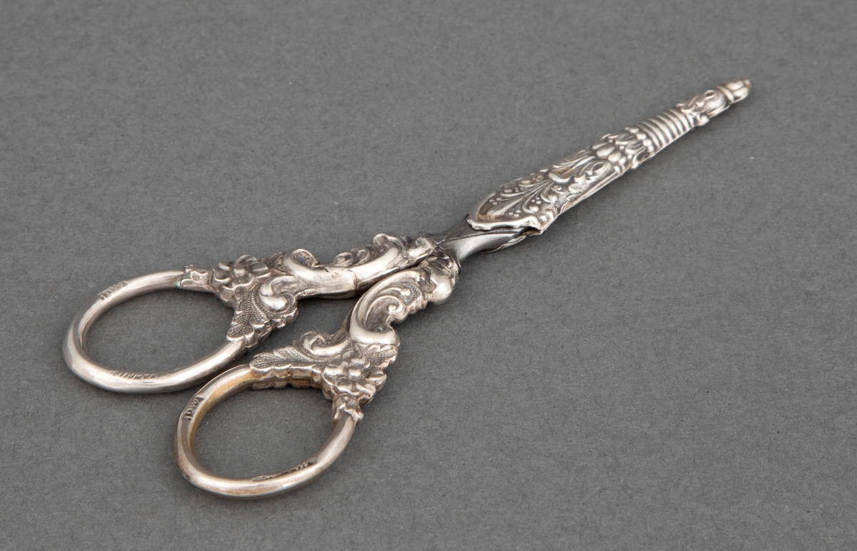 Brodersaks av stål med etsede ornamenter; blomster, ranker og palmetter og ornamentikk i presset sølvblikk med blomster og ranker i nyrokokko. Til saksen hører en liten slire av presset og mønstret sølvblikk.