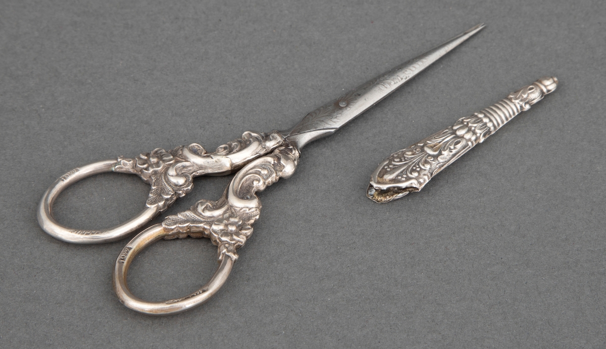 Brodersaks av stål med etsede ornamenter; blomster, ranker og palmetter og ornamentikk i presset sølvblikk med blomster og ranker i nyrokokko. Til saksen hører en liten slire av presset og mønstret sølvblikk.