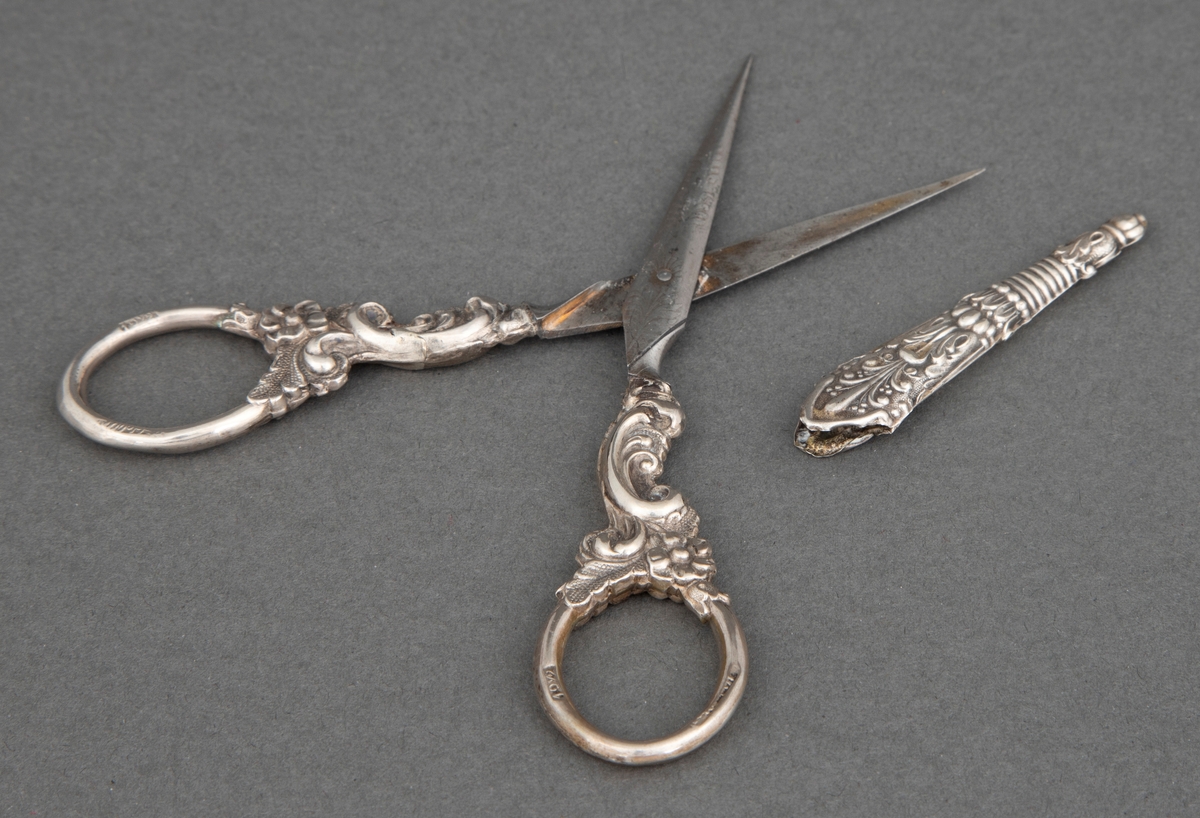 Brodersaks av stål med etsede ornamenter; blomster, ranker og palmetter og ornamentikk i presset sølvblikk med blomster og ranker i nyrokokko. Til saksen hører en liten slire av presset og mønstret sølvblikk.