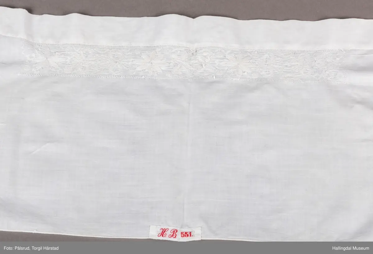 Den ene langsides midtparti har innefor bred fald, bord av 3 rosetter og 7 sløyfelignende ornamenter. Kortsidene er kantet med knipling og prydet med et bredt parti av ruter i hardangersøm. Hodkluten er ved tverrsømmer sammensatt av 5 forskjellige deler. Endestykkene med hardangersømmen er av grovere tøy enn det øvrige. 
 R.B.S.