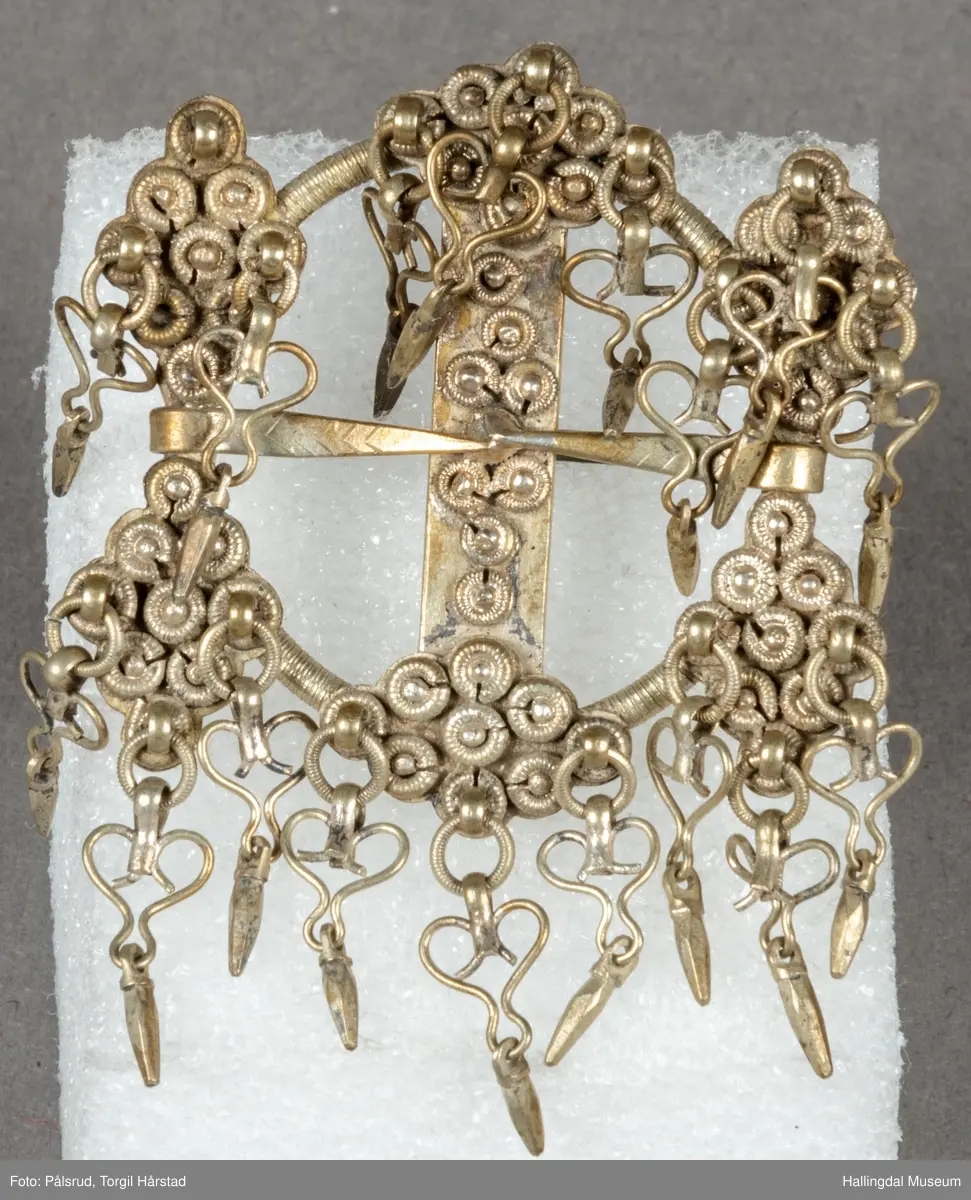 Byggkornsølje, sølv. Ring med tverribbe og 2 nåler. Øverst og nederst på ribben samt over og under begge nåler rombeformet plate dekket av 9 kuler med riftlet rand, hver rombe har 3 byggkorn, dobber. De 2 nåler danner tilsammen en rett linje. Dobbene består av en herteformet bøyle med en løkke nederst hvori der henger en liten tapp.
R.B.S.