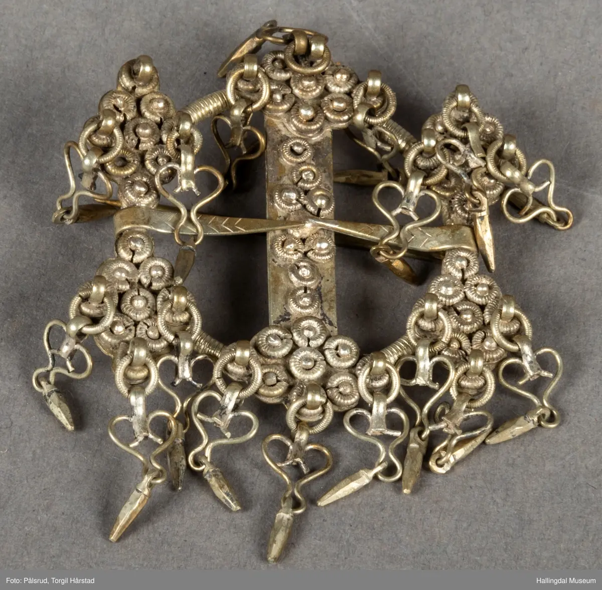 Byggkornsølje, sølv. Ring med tverribbe og 2 nåler. Øverst og nederst på ribben samt over og under begge nåler rombeformet plate dekket av 9 kuler med riftlet rand, hver rombe har 3 byggkorn, dobber. De 2 nåler danner tilsammen en rett linje. Dobbene består av en herteformet bøyle med en løkke nederst hvori der henger en liten tapp.
R.B.S.