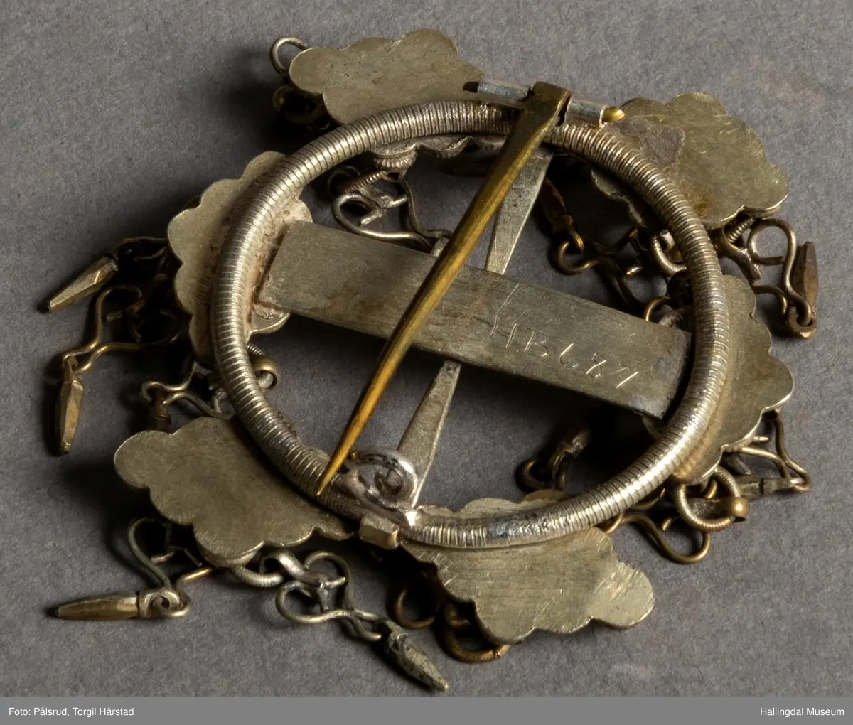 Byggkornsølje, sølv. Ring med tverribbe og 2 nåler. Øverst og nederst på ribben samt over og under begge nåler rombeformet plate dekket av 9 kuler med riftlet rand, hver rombe har 3 byggkorn, dobber. De 2 nåler danner tilsammen en rett linje. Dobbene består av en herteformet bøyle med en løkke nederst hvori der henger en liten tapp.
R.B.S.