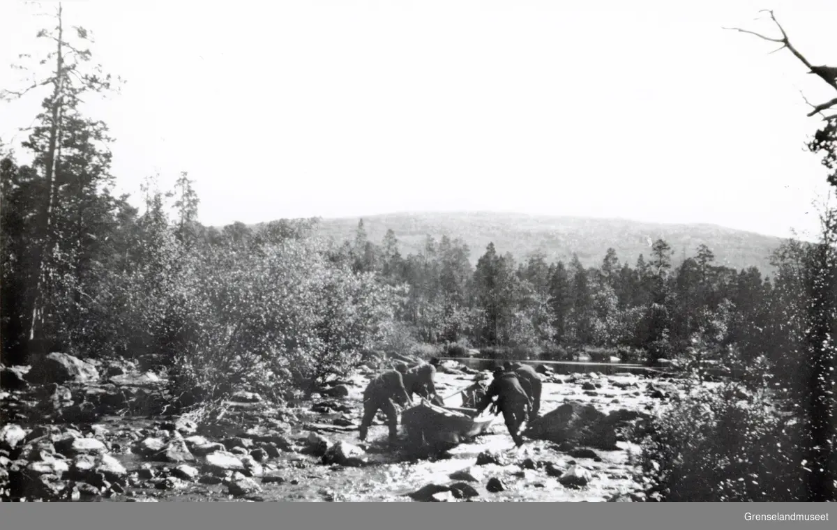 Motstrøms ferdsel med båt i Kivilombola (steinstrykene), ved grensa mellom Norge og Finland i Pasvikdalen. 2 i Finland og 3 i Norge. Båten trekkes oppover i midtålen av fem mann. Bjørke- og furuskog på elvebreddene. 
