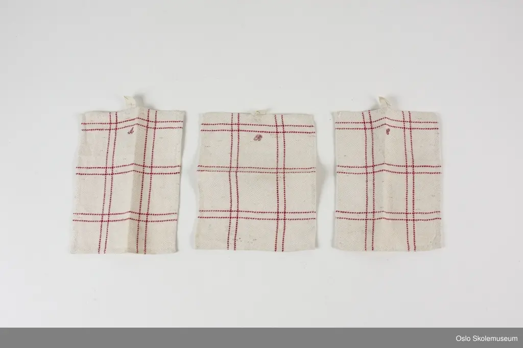 Tre håndklær miniformat med hempe. Håndklærne er hvite/beige med røde sting i et stripemønster. De tre håndklærne er merket med hver sin broderte bokstav; "A", "B" og "C".