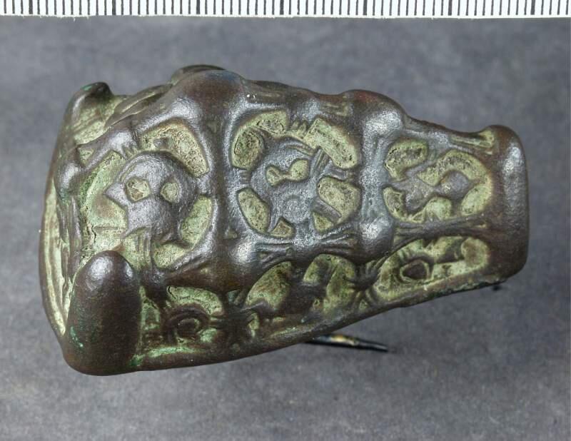 Spänne med ornamentik i kraftig relief, ovansidan indelad i fält med vulstförsedda lister, bottenplatta och nål bevarade, ganska nött
av brons, djurhuvudformat, längd 59, br. 36 h. 30 mm. 3 i längd - och 3 i tvåriktning gående med hög ornering prydda band, mellan dem kraftig ornering, (nålspetshylsan saknas). Förut märkt: 229.
